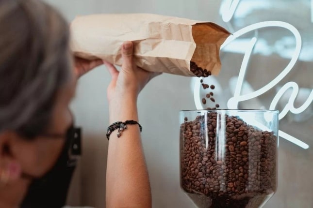 Mulher branca de cabelos grisalhos segurando um saco de café de papel pardo, despejando os grãos em um recipiente de vidro quase cheio