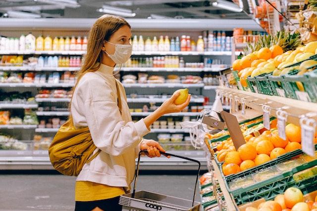 Mulher branca de cabelos longos, loiros e lisos, em um supermercado, segurando um limão siciliano na mão e usando máscara