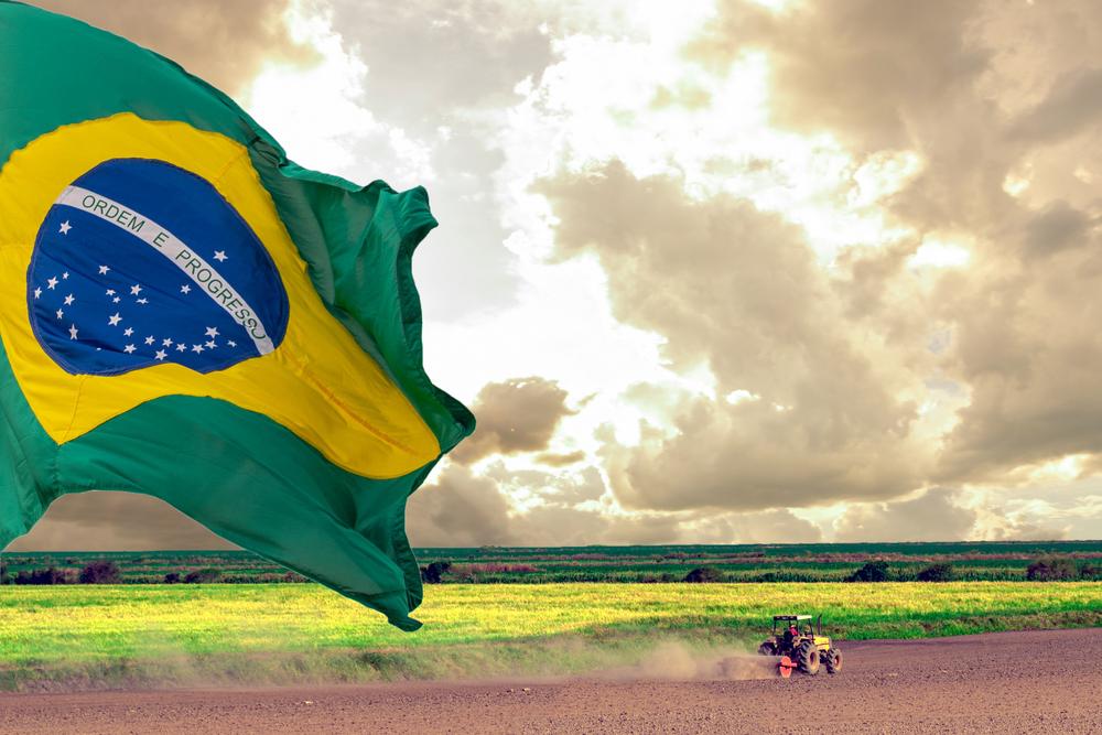 Céu nublado com bastante nuvens, campo verde ao fundo e trator andando na terra, com bandeira do Brasil no canto esquerdo da imagem