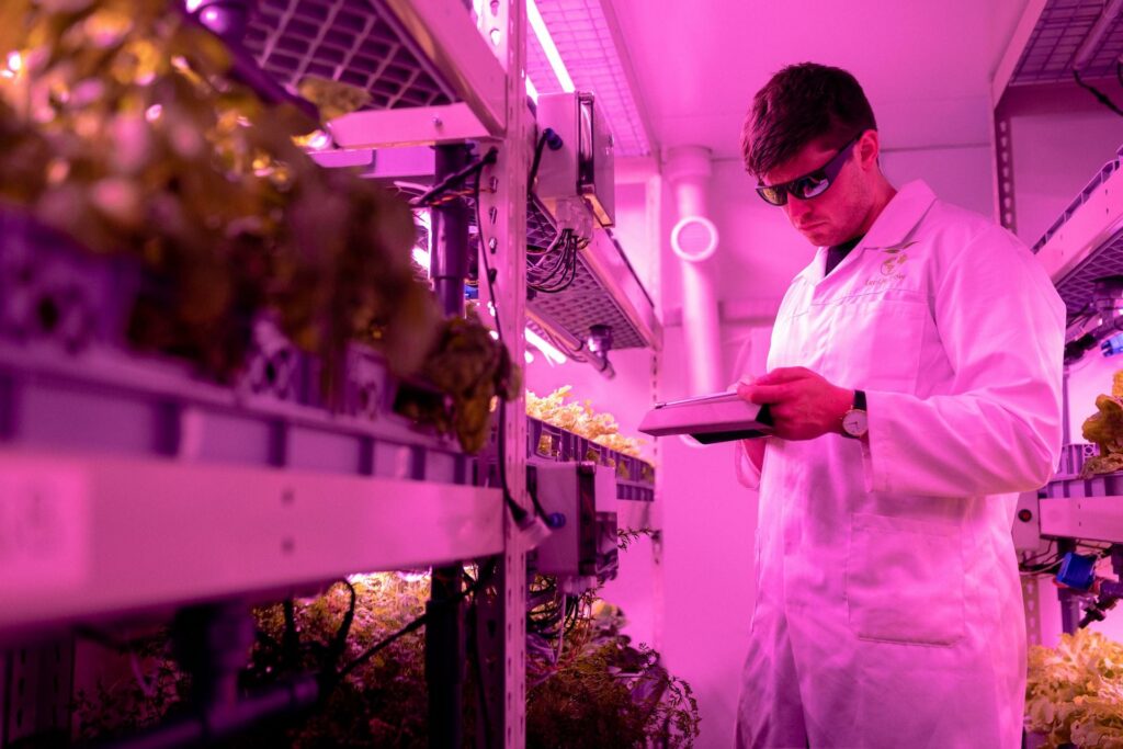 Homem branco de cabelo preto curto, vestido com jaleco branco, em um laboratório com mudas de plantas e luz rosa